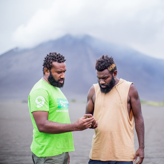 Portrait of two men standing outside. One is holding a phone while the other, wearing a lime green Oxfam t-shirt is as well. Yasur volcano is in the background.