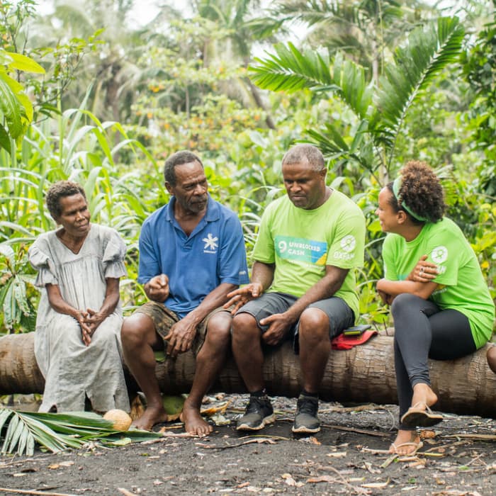 Four people are sitting on a log outside in a forest. Two of them are wearing green Oxfam Australia t-shirts.