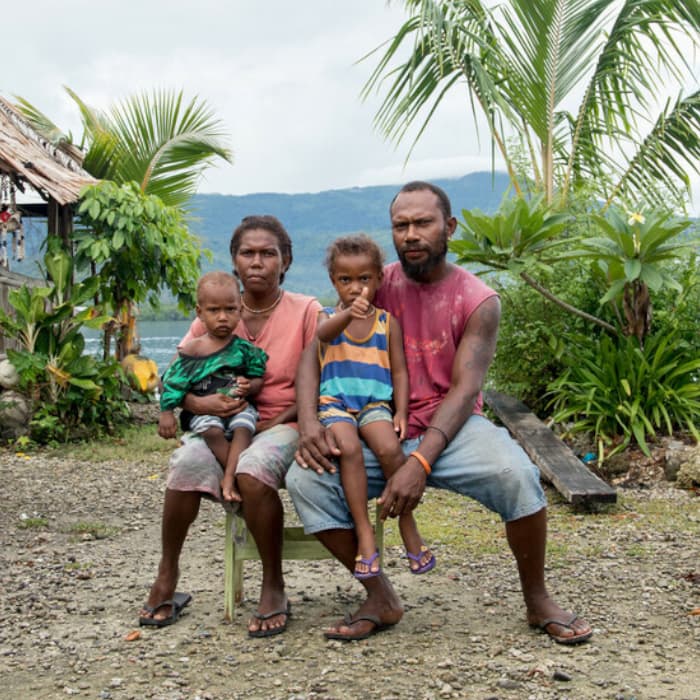 A family is sitting on a single chair outside of their home on Loreto Island, Malaita province. They have two young children and one is holding a thumbs up gesture.