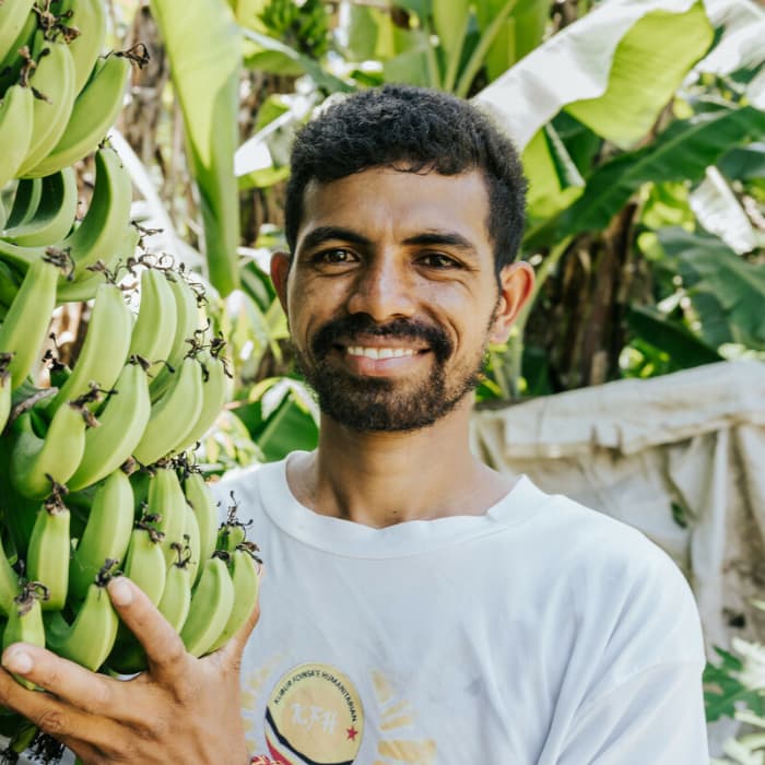 A closeup of a smiling man. He is holding up a bunch of green bananas.