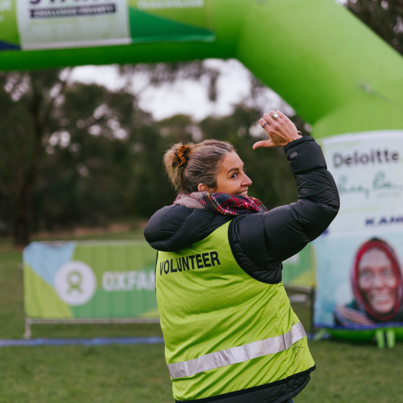 A woman is showing off the back of her green vest that reads "Volunteer". She is standing outdoors at the Melbourne Trailwalker 2022 event.